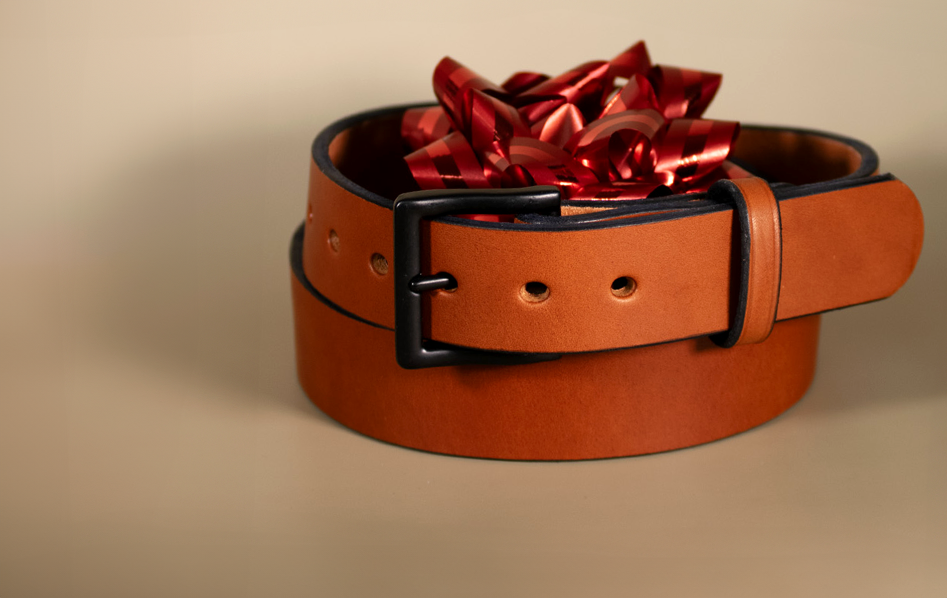 A brown leather belt coiled into a circle with a black buckle, topped with a shiny red gift bow, placed on a neutral background.