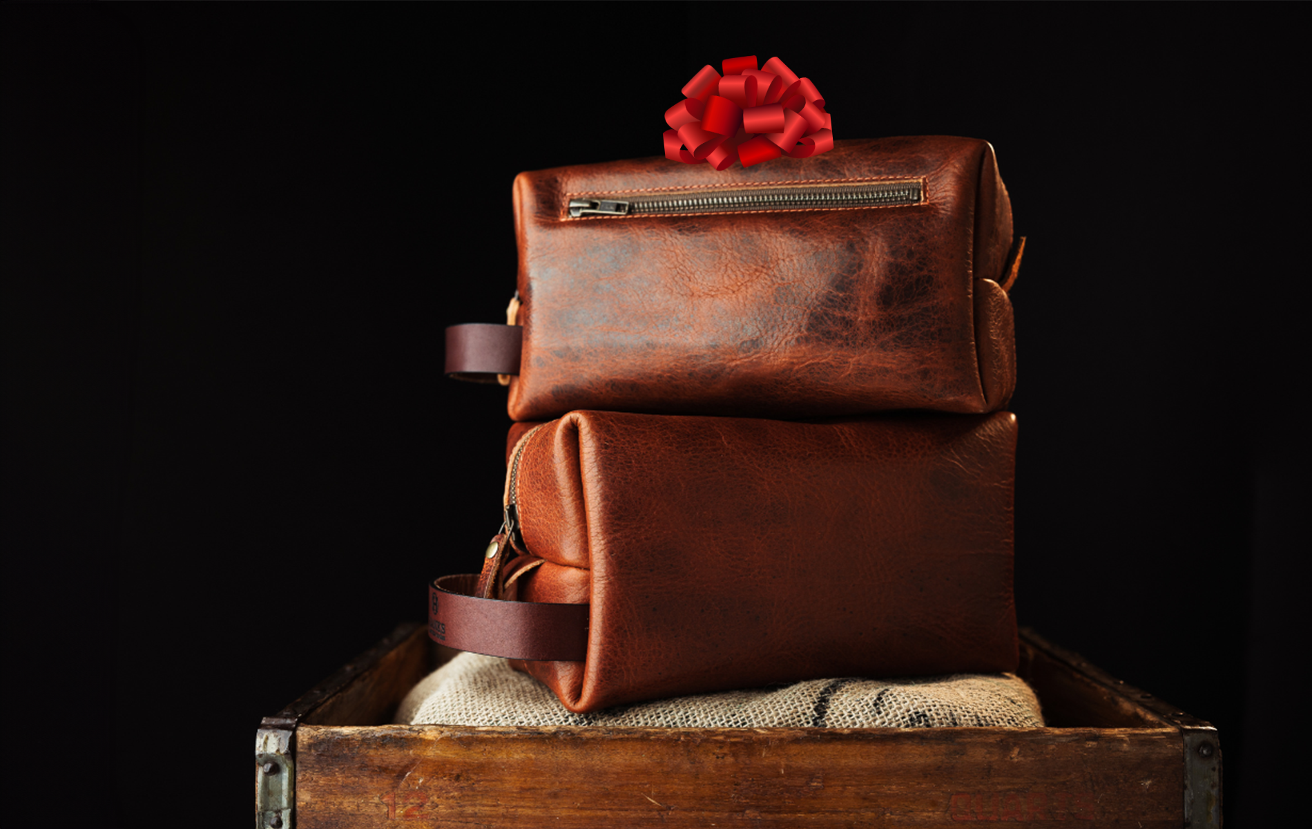 Two stacked brown leather bags sit on a wooden box against a dark background. The top bag is adorned with a red bow, suggesting a gift.