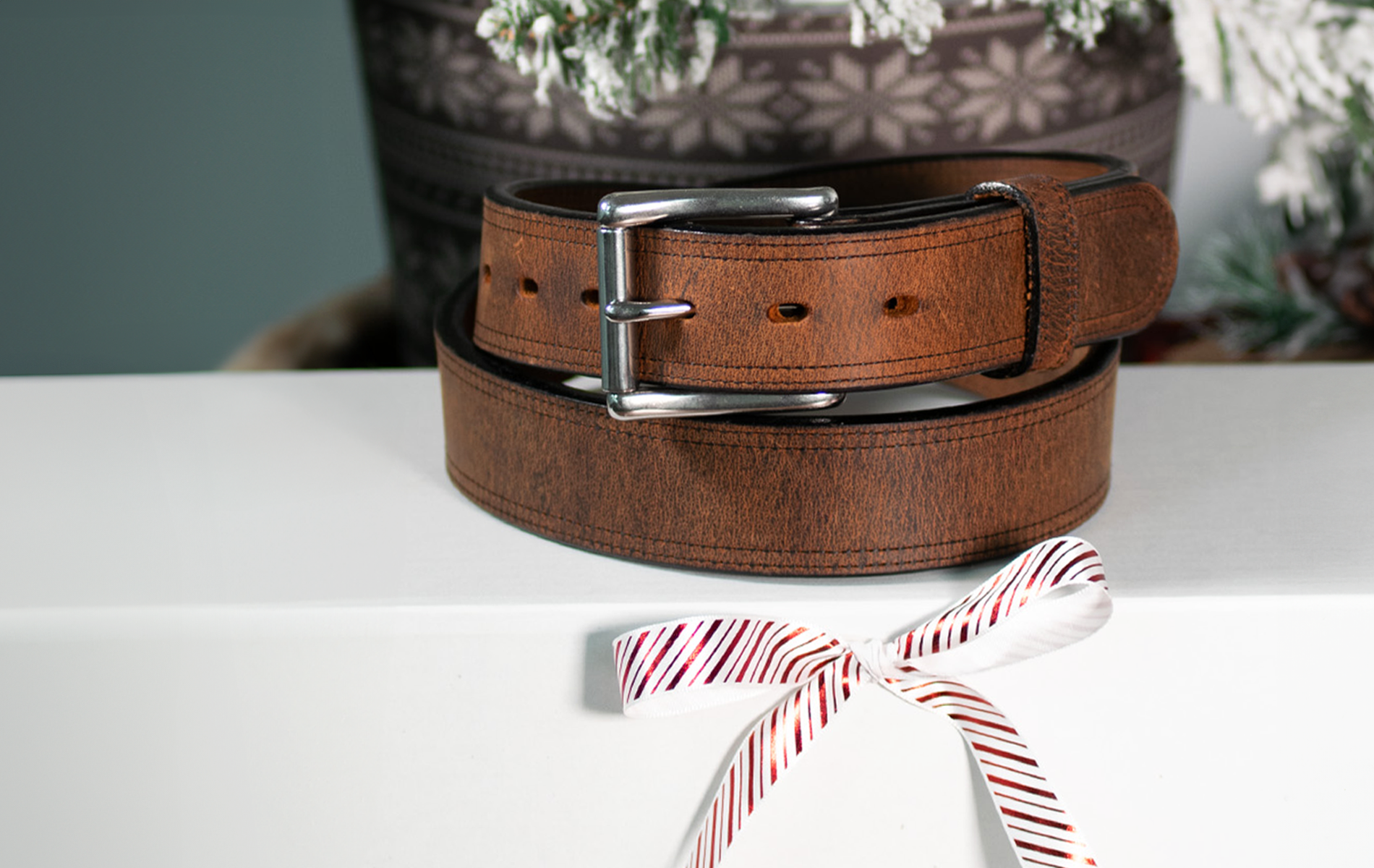 A brown leather belt with a silver buckle is placed on a white surface, decorated with a red and white striped ribbon. In the background, a patterned, snowflake-themed object is partially visible.