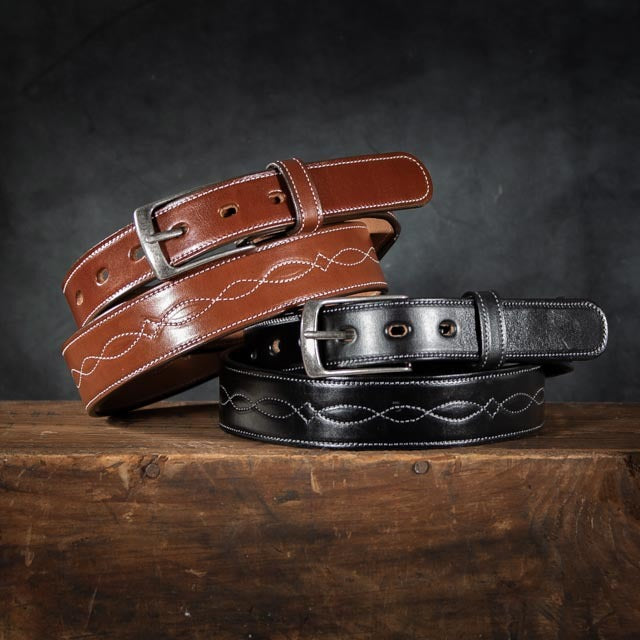 Three handcrafted leather belts on a wooden surface against a dark background include the top Teton belt from Hanks Belts, which is brown with a silver buckle, and the bottom belt made from Premium English Bridle Vegetable Tanned Leather with white stitching and a sleek black buckle.