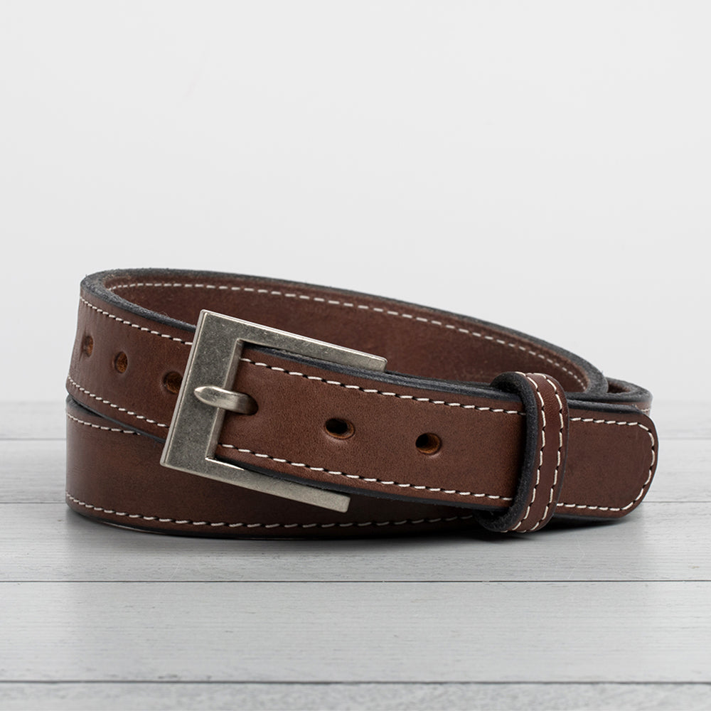 The Bonnie Belt - 1.25&quot; from Hanks Belts, made of full-grain leather with white stitching and a sleek silver rectangular buckle, is coiled neatly on a light gray wooden surface.