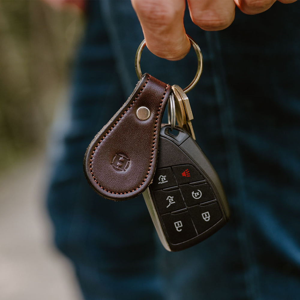 A hand holds a set of car keys attached to the Hanks Havana Bell-Shaped Leather Keychain, crafted from full grain leather. The key fob features several buttons, including lock, unlock, and alarm. The background is blurred, emphasizing the keys and the stylish keychain.