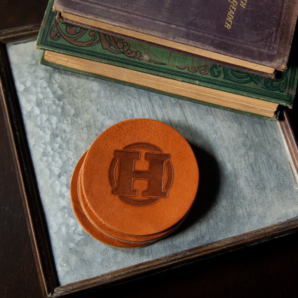 A stack of Round Leather Coasters - Set of 4 from Hanks Belts, crafted from premium full grain leather with an embossed &quot;H&quot; logo, sits on a textured metal surface. Made in the USA, these coasters rest atop two closed books with worn covers, one green and the other purple, adding a touch of elegance to the scene.