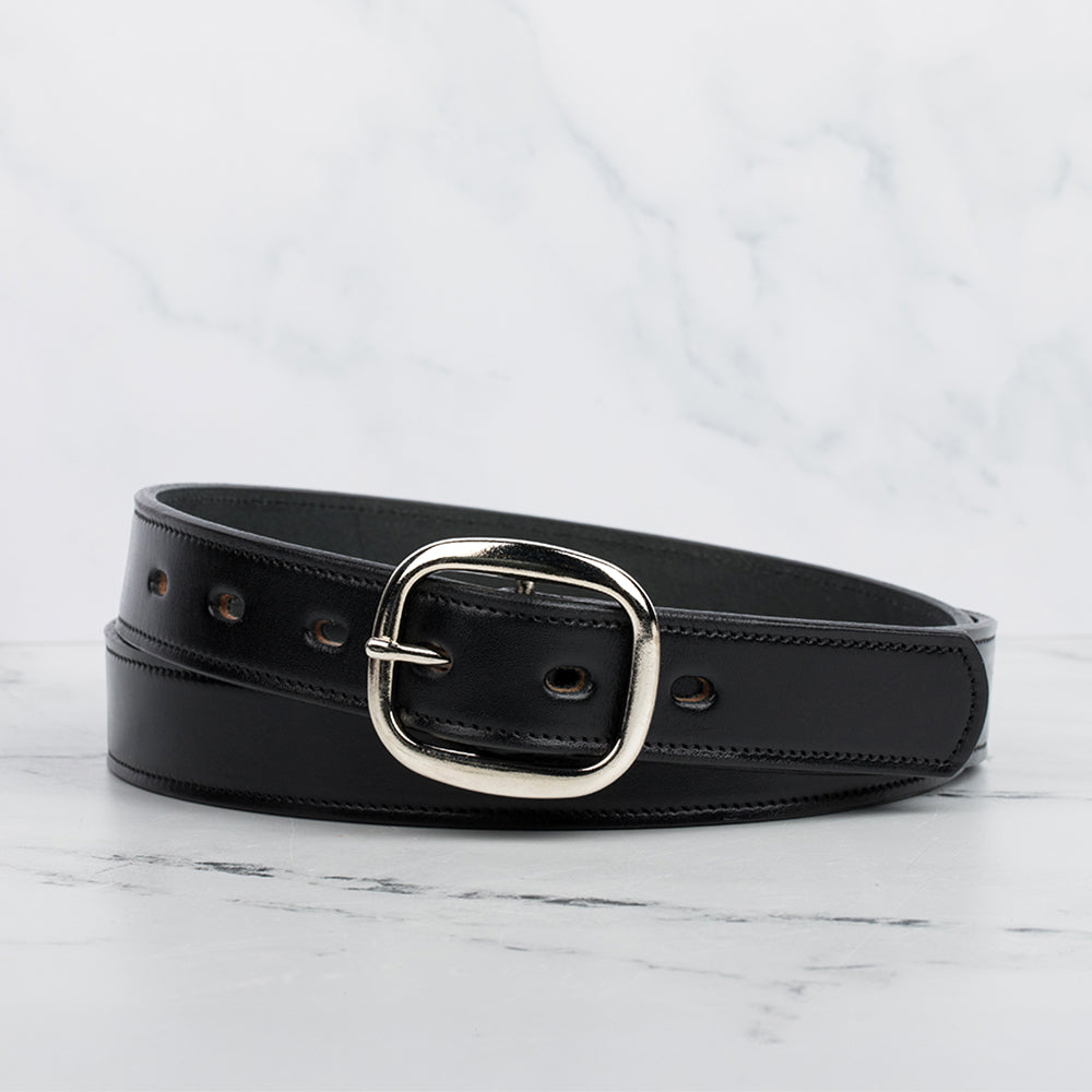 The black Eloise Belt - 1.25&quot; by Hanks Belts, crafted from premium vegetable-tanned leather, features a shiny silver center bar buckle and several adjustment holes. It is coiled neatly on a light marble surface.