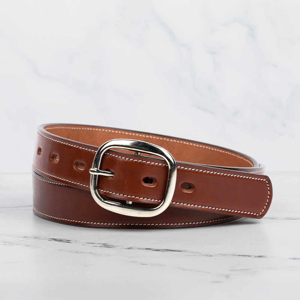 The Eloise Belt - 1.25&quot; by Hanks Belts is a women&#39;s premium accessory made of brown vegetable-tanned leather with white stitching, featuring a shiny silver oval buckle. It lies on a white marble surface, coiled in a loose spiral with evenly spaced holes along its length.