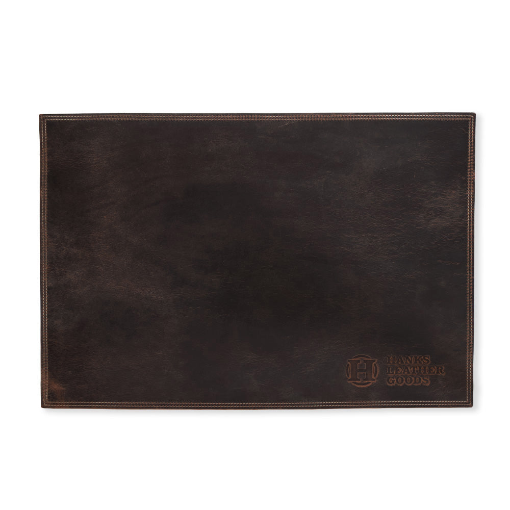 A rectangular desk pad crafted from premium dark brown bison leather, showcasing visible stitching and an embossed logo in the bottom right corner reading "Hanks Belts." Handmade in the USA, its surface is smooth with a slightly worn appearance.