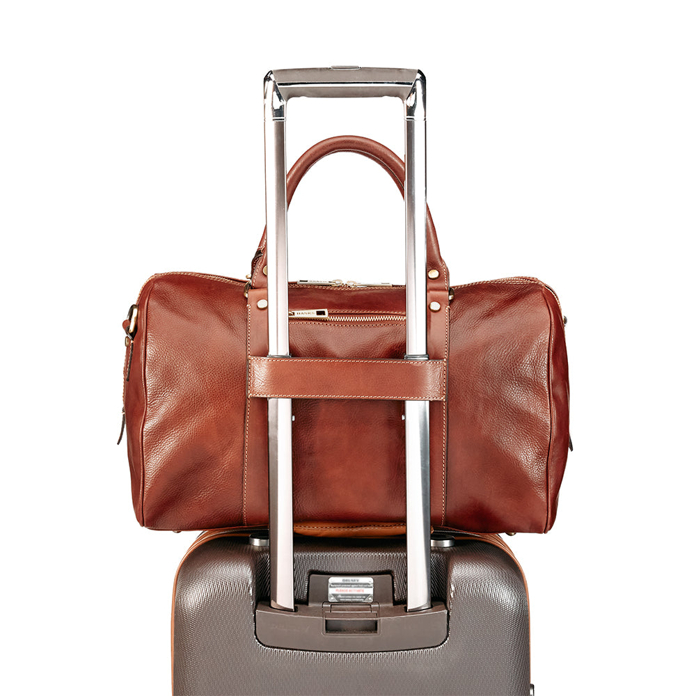 The Getaway Duffel With Luggage Sleeve by Hanks Belts, crafted in brown Italian leather, is stylishly affixed to the extended handle of a silver hard-shell suitcase. This combination captures the essence of luxury travel against a clean, minimalist backdrop, highlighting its carry-on-friendly design.