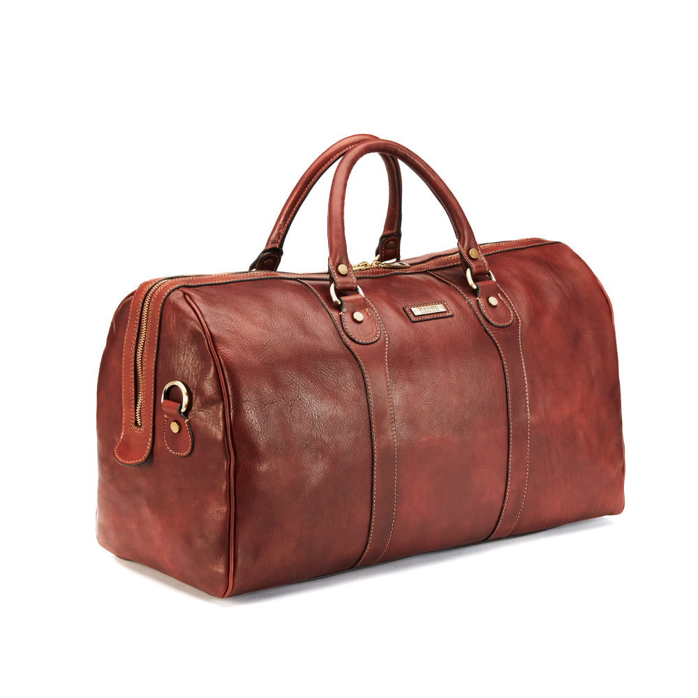 The Wanderer Duffel by Hanks is an Italian-made brown leather bag featuring sturdy handles, metal accents, and a zipper closure. This luxury piece offers a smooth finish and an elegant design, showcased against a plain white background at an angle.