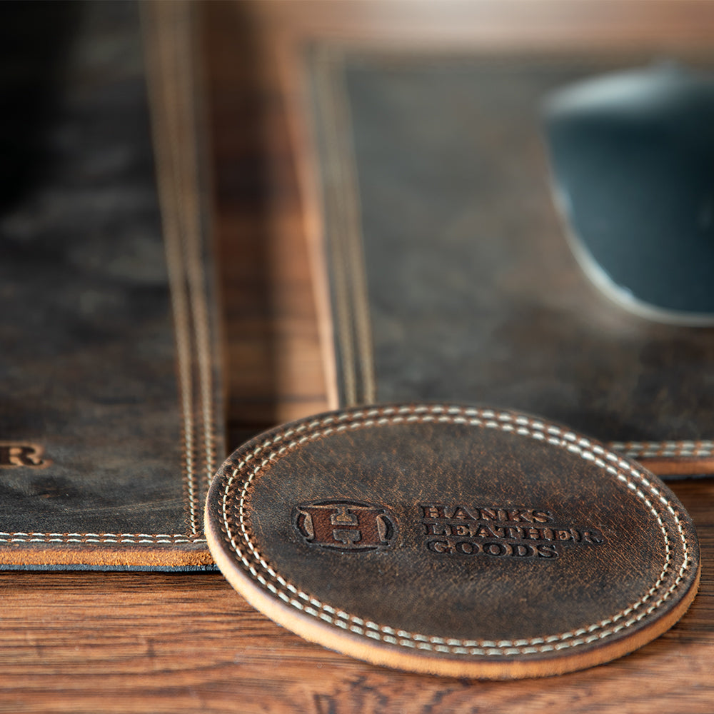 Close-up of a Premium Round Leather Coaster from Hanks Belts on a wooden surface. Part of a leather desk set, the coasters come with visible stitching that suggests a rustic, handcrafted aesthetic and includes a matching mat.