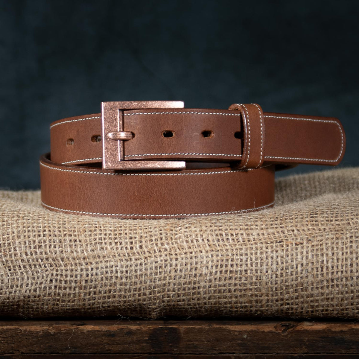 The Heritage - 1.25&quot; from Hanks Belts is elegantly coiled and displayed on a piece of burlap fabric against a dark background, exuding rugged appeal. Made from bison leather, this belt features white stitching along its edges and a rectangular metal buckle, showcasing American craftsmanship.