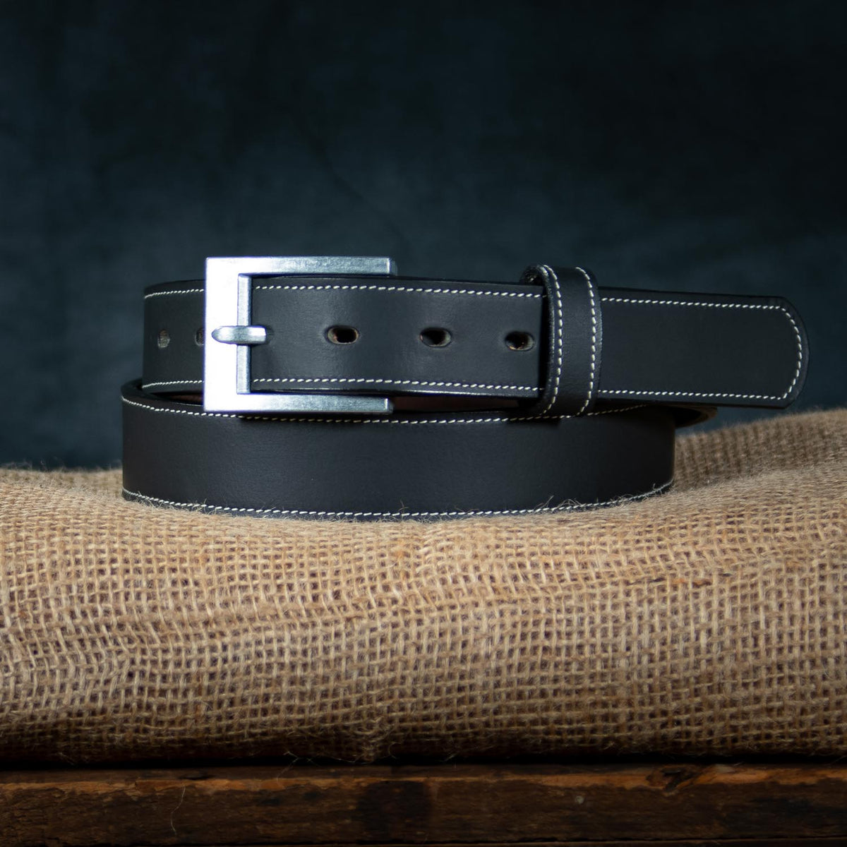 Coiled on a piece of burlap is The Heritage - 1.25&quot; by Hanks Belts, a black leather belt with white stitching. This rugged bison leather belt features a silver rectangular buckle and multiple holes for adjustment. The dark background enhances the contrast with the light-colored burlap, accentuating the belt&#39;s rugged appeal.