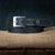 Coiled on a piece of burlap is The Heritage - 1.25" by Hanks Belts, a black leather belt with white stitching. This rugged bison leather belt features a silver rectangular buckle and multiple holes for adjustment. The dark background enhances the contrast with the light-colored burlap, accentuating the belt's rugged appeal.