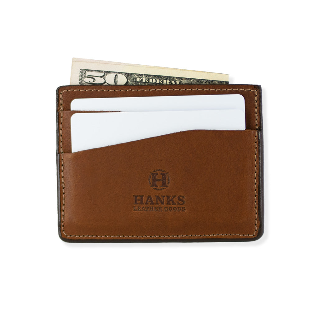 Introducing the Cumberland Bison Card Holder by Hanks Belts: a peanut brown leather card holder crafted from durable Bison Leather, showcasing the brand name "Hanks Leather Goods" with an embossed logo on the front. This ultra-thin, lightweight wallet is made in the USA and features two slanted card slots along with visible stitching along the edges for a timeless look.