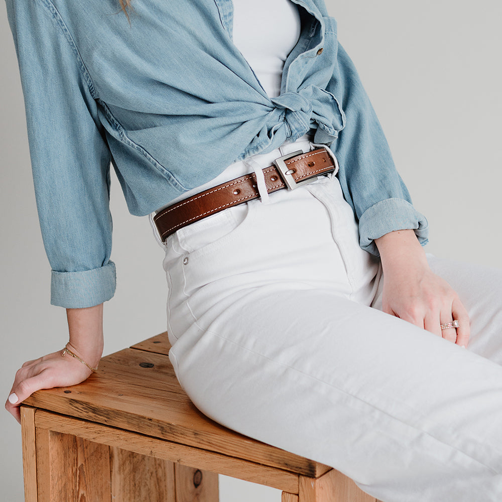 A person sits on a wooden stool, wearing a light blue button-up shirt tied at the waist, white high-waisted pants, and a USA-made Hanks Belts Bonnie Belt - 1.25&quot; crafted from full-grain vegetable-tanned leather. Their left hand rests on the stool, displaying a ring on their finger.