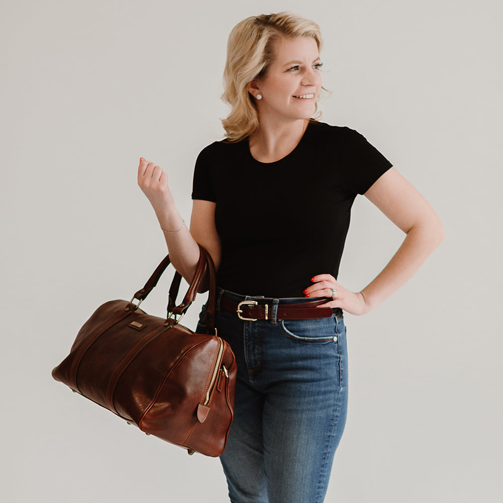 A blonde woman in a black t-shirt and jeans holds the Hanks Belts&#39; Getaway Duffel With Luggage Sleeve, made from full-grain Italian leather. She stands against a light background, smiling and looking to the side, exemplifying an ideal travel companion.