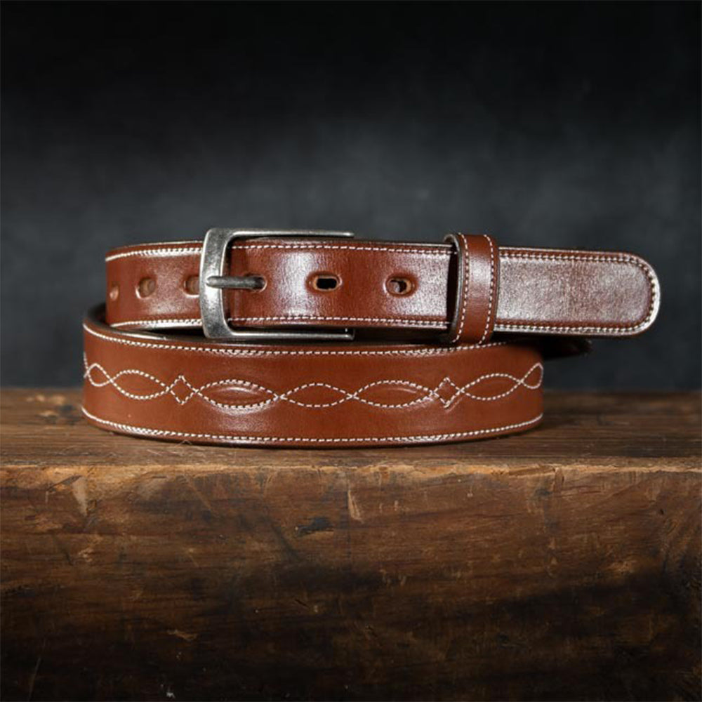 The Teton - 1.5&quot; by Hanks Belts is a premium leather belt featuring a silver buckle and decorative white stitching in a looping pattern, elegantly handcrafted and displayed coiled on a wooden surface against a dark background.