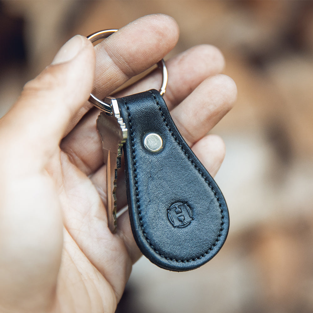 In a softly blurred background emphasizing its elegant craftsmanship, a hand holds the Black Bell-Shaped Leather Keychain from Hanks. Crafted from full grain leather, it features a metal rivet  and an embossed &quot;H&quot; logo, with a single key attached to the keyring.