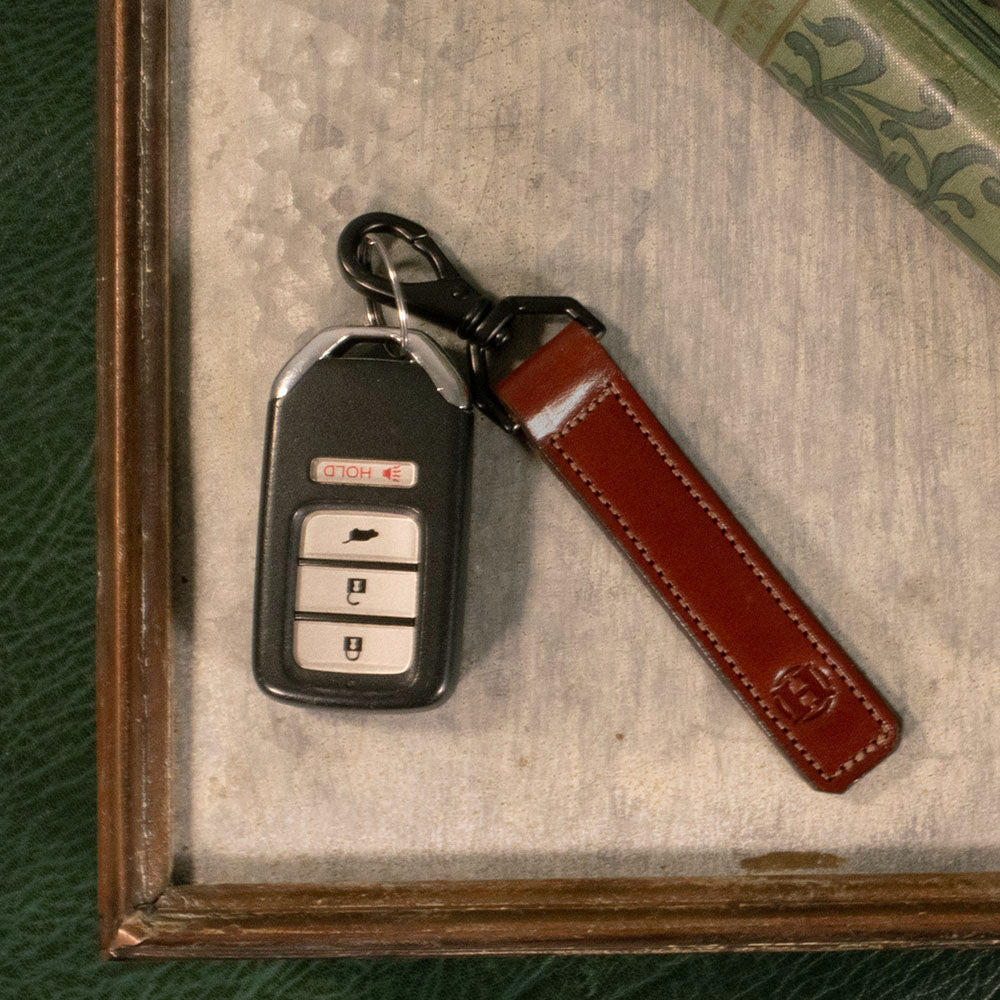 An Effortless Keychain by Hanks Belts, featuring a car key fob with three buttons (lock, unlock, and trunk) and crafted from USA-tanned vegetable tanned full grain leather, rests on a textured, light-colored surface next to a green patterned fabric.