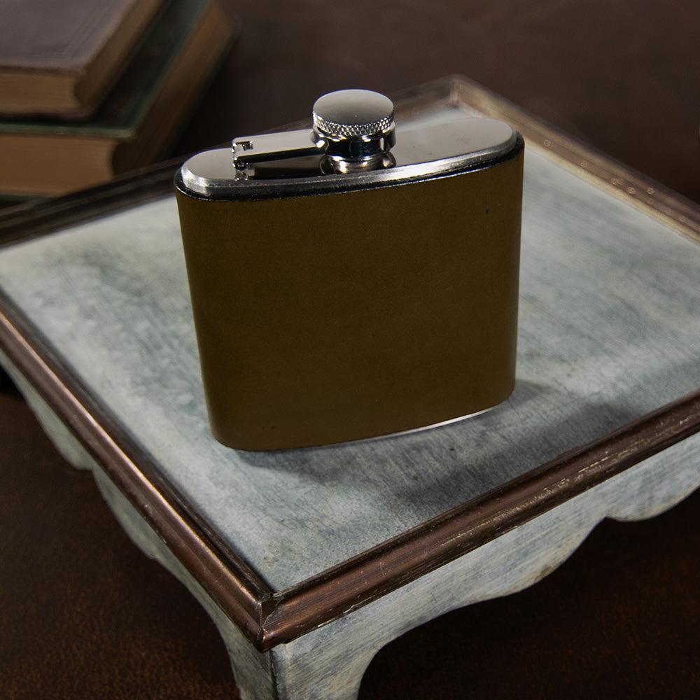 A Hanks Belts Leather Covered Flask sits on a small, vintage, distressed wood tray. The tray is placed on a rustic surface, with a stack of old books blurred in the background, creating a nostalgic and classy atmosphere.