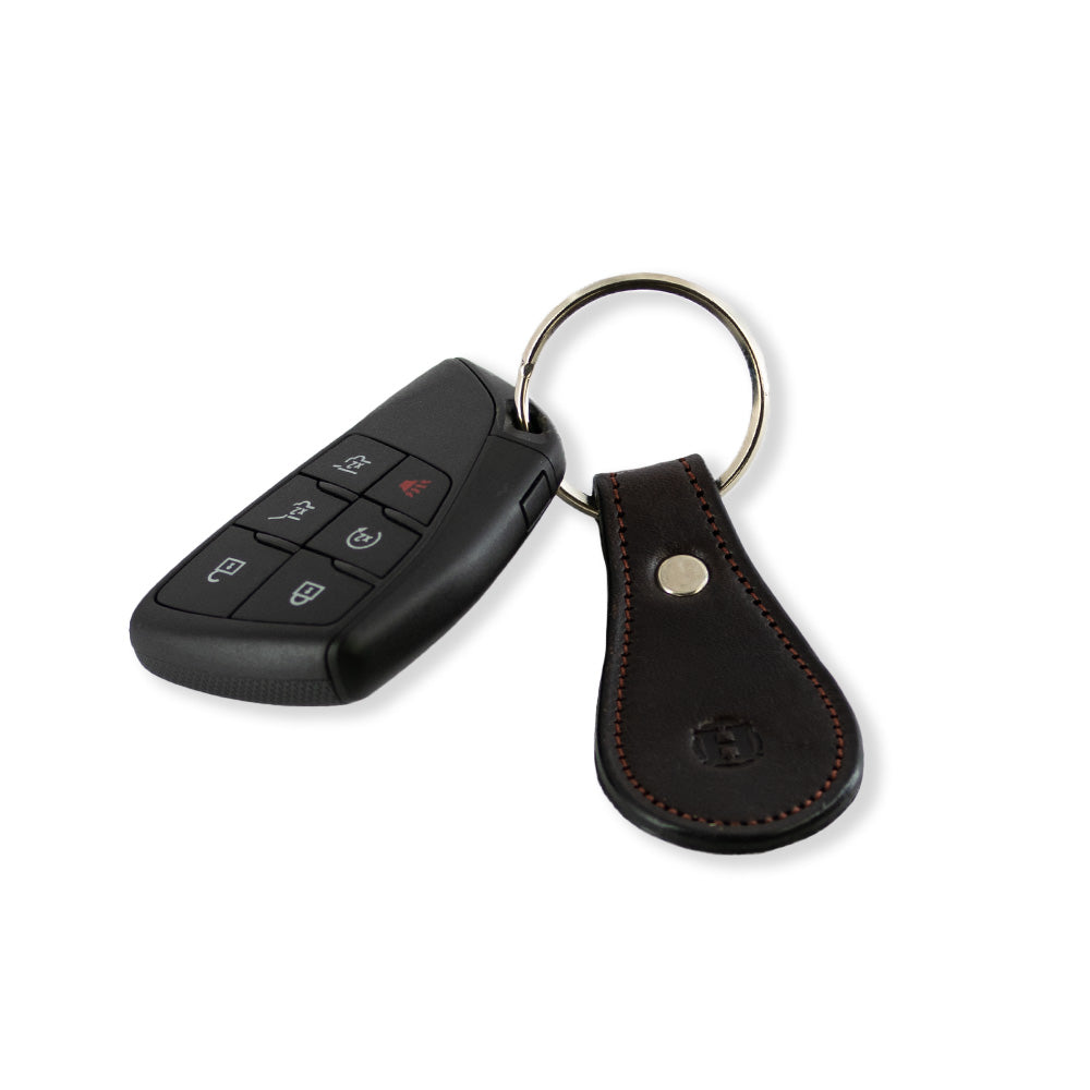 A black car key fob with multiple buttons, including lock, unlock, and panic, is attached to Hanks Belts&#39; Bell-Shaped Leather Keychain in Havana full grain leather with a metal ring and decorative stitching detail. Made in the USA.