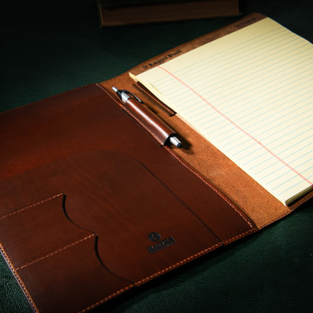 A leather portfolio open on a dark surface. The left side features a pocket with small slots for cards and a pen holder, holding a silver pen. Crafted from full-grain leather, the right side includes a yellow lined notepad. The USA-made Luxury Folio by Hanks Belts proudly displays the &quot;Albert Tusk&quot; logo embossed on the leather.