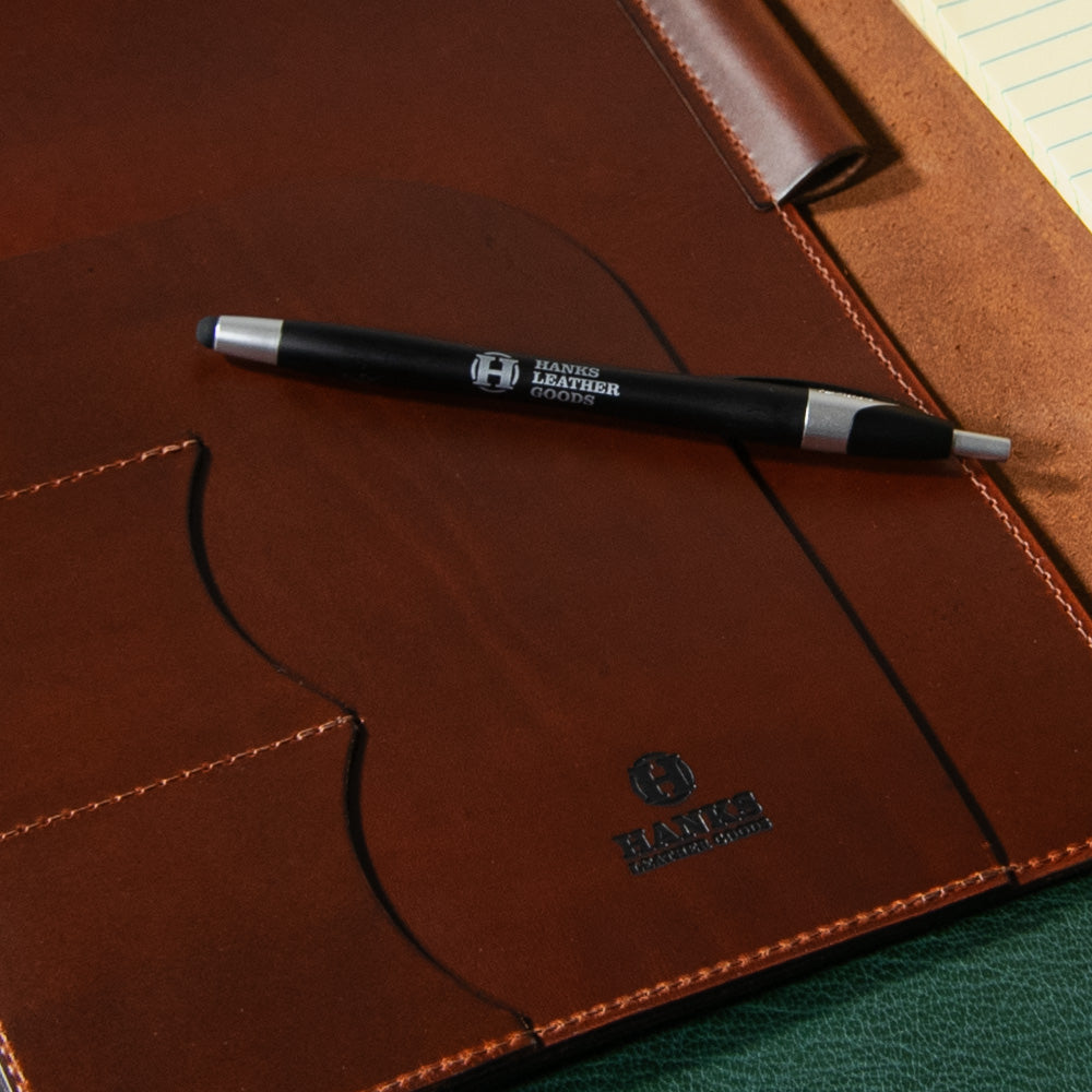 A full-grain leather luxury folio with a pen on top is shown. The portfolio, named &quot;The Luxury Folio,&quot; is brown with visible stitching and the logo &quot;HAK&quot; along with text underneath. The black pen has branding that reads &quot;Hanks Belts.&quot; A portion of a lined notepad can be seen in the corner, exemplifying superior USA made craftsmanship.