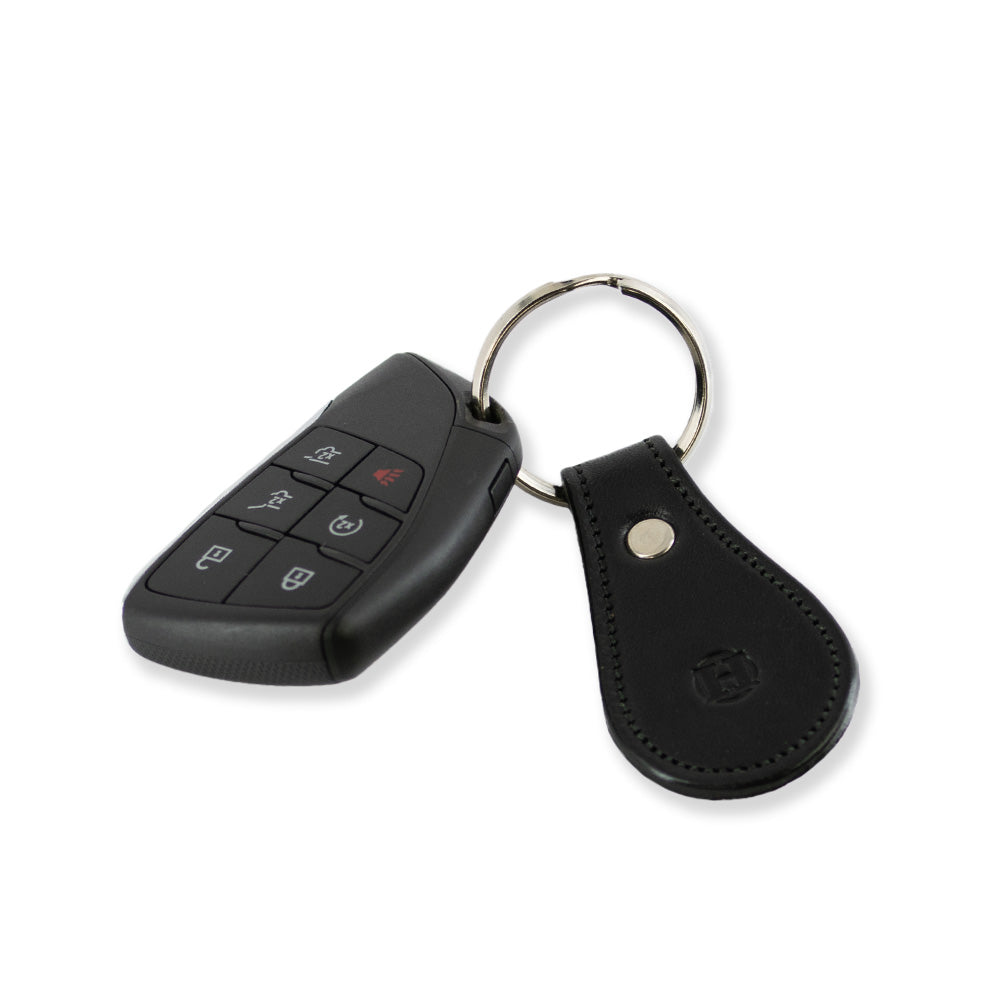 A black electronic car key fob with four buttons and a red alert button is attached to a key ring. The key ring also holds a Hanks Belts Black Bell-Shaped Leather Keychain, made in the USA, featuring a metal stud and a subtle embossed logo. The background is plain white.