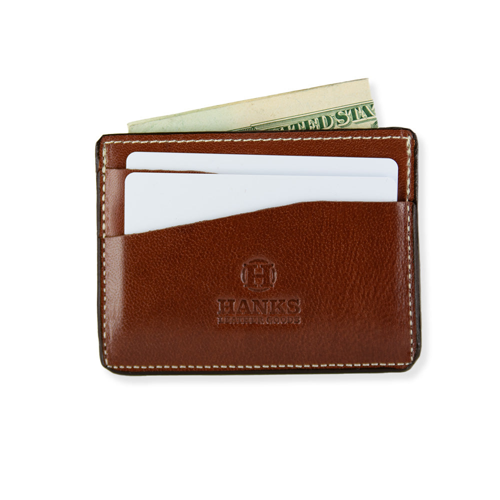 A brown Monroe Goatskin Card Holder by Hanks featuring a visible brand logo &quot;Hanks Leather Goods&quot; embossed on the front. This minimalist wallet contains two credit cards and a partially visible folded U.S. dollar bill, showcasing its slim profile and functional design with multiple card and currency slots against a white background.