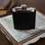 A Hanks Belts Leather Covered Flask with a metallic cap is placed on a square, weathered wooden tray. The tray sits on a dark brown surface, and a couple of closed books are partially visible in the background, enhancing the vintage feel of the overall setting.