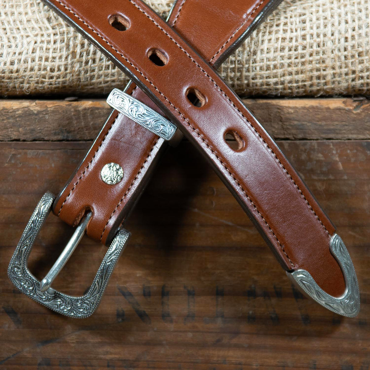 A brown leather belt with intricate silver buckles and an embroidered Aztec pattern is displayed against a rustic wooden surface and burlap backdrop. The Seneca - 1.5&quot; by Hanks Belts, made from premium English bridle vegetable-tanned leather, is laid out in an X shape, showcasing both the buckle and the punch holes.