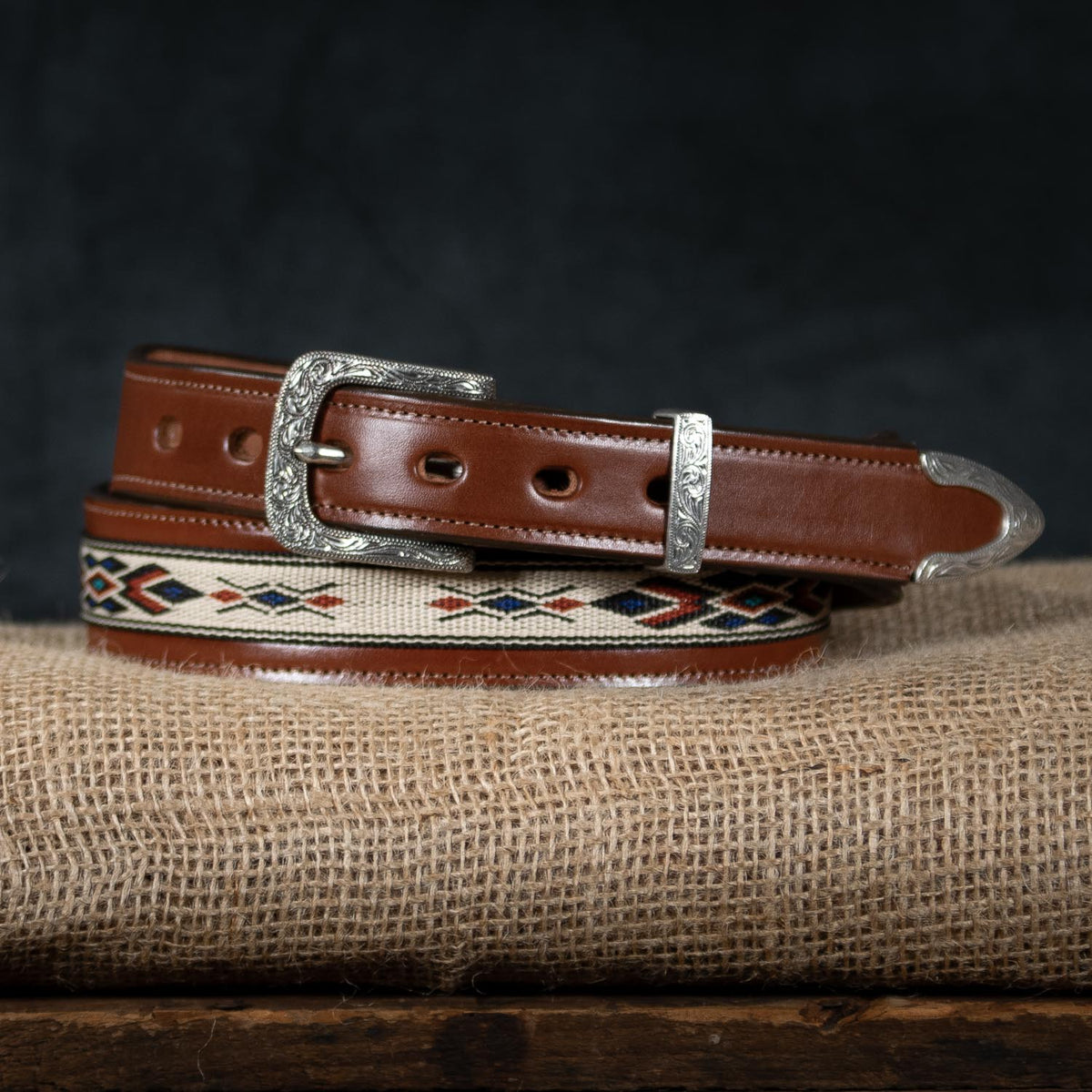 The Seneca - 1.5&quot; by Hanks Belts is a brown leather belt with a silver buckle and end tip, adorned with an intricate embroidered Aztec pattern in red, black, and white. Made from Premium English Bridle Vegetable-Tanned Leather, the belt is rolled up and showcased on burlap fabric against a dark background.