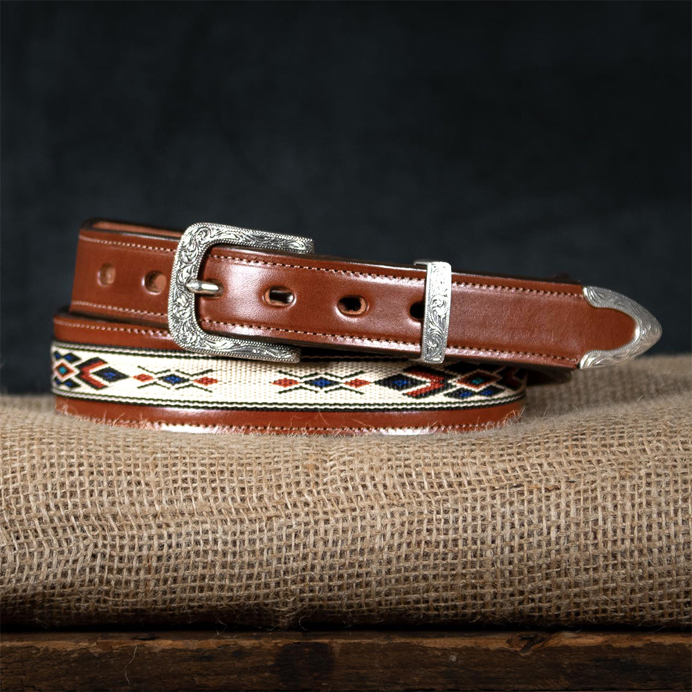 The Seneca - 1.5&quot; by Hanks Belts, crafted from premium English bridle leather, features decorative red, blue, and beige stitching. It includes an ornate silver buckle and tip, exuding a western spirit as it sits coiled on a burlap surface.