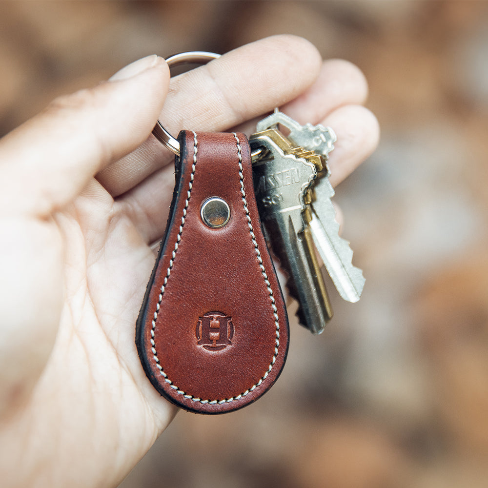 A hand holds the Oak Bell-Shaped Leather Keychain from Hanks Belts, featuring a circular embossed logo on full-grain leather with edge stitching.