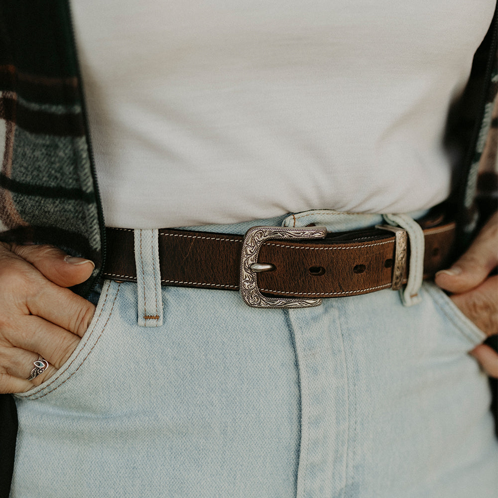 A person wearing light blue jeans with hands in pockets and a women&#39;s western belt from Hanks Belts, featuring the ornate floral buckle set of the Willow Belt - 1.25&quot;. The ensemble also includes a white shirt partially covered by a plaid jacket, highlighting both style and durability.