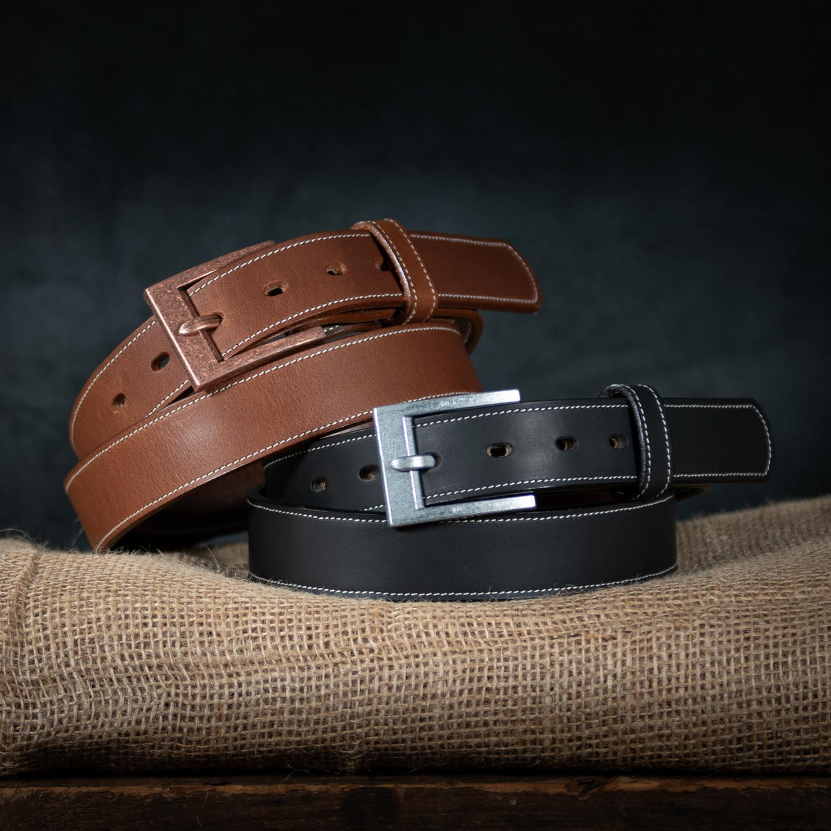 Two leather belts from Hanks Belts&#39; The Heritage - 1.25&quot; collection, one brown and one black, are displayed on a piece of burlap fabric. The brown belt features a brass buckle, while the black belt sports a silver buckle. Both belts boast white stitching along the edges, adding a touch of rugged appeal to their design and showcasing American craftsmanship.