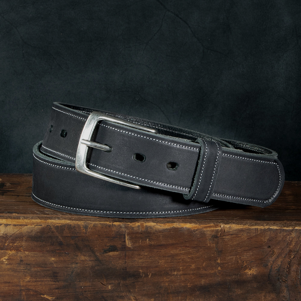 The Suede Belt - 1.5&quot; by Hanks Belts, crafted from USA-made materials, features a silver buckle and double stitching. It is coiled on a wooden surface against a dark background, showcasing its multiple holes.