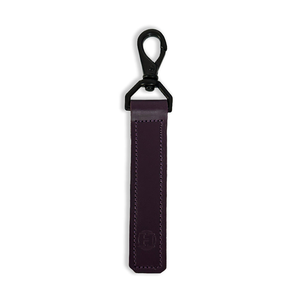 The Effortless Keychain by Hanks Belts is crafted from USA Tanned Vegetable Tanned Full Grain Hides in a dark purple color. It features black stitching, a metal clip for easy attachment to keys or bags, and an embossed logo at the bottom. The design is both sleek and minimalist.