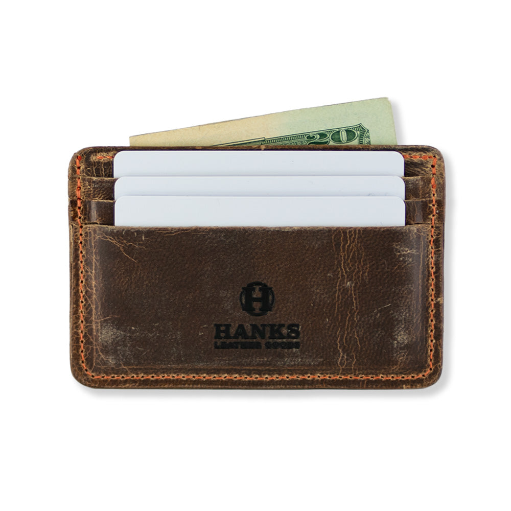 The Hamlin Card Holder with ID Window by Hanks Belts is a brown, worn-looking accessory made from durable Steerhide leather. It features three slots on the front and stitching around the edges. Proudly made in the USA, it showcases the "HANKS" logo in black at the bottom center.