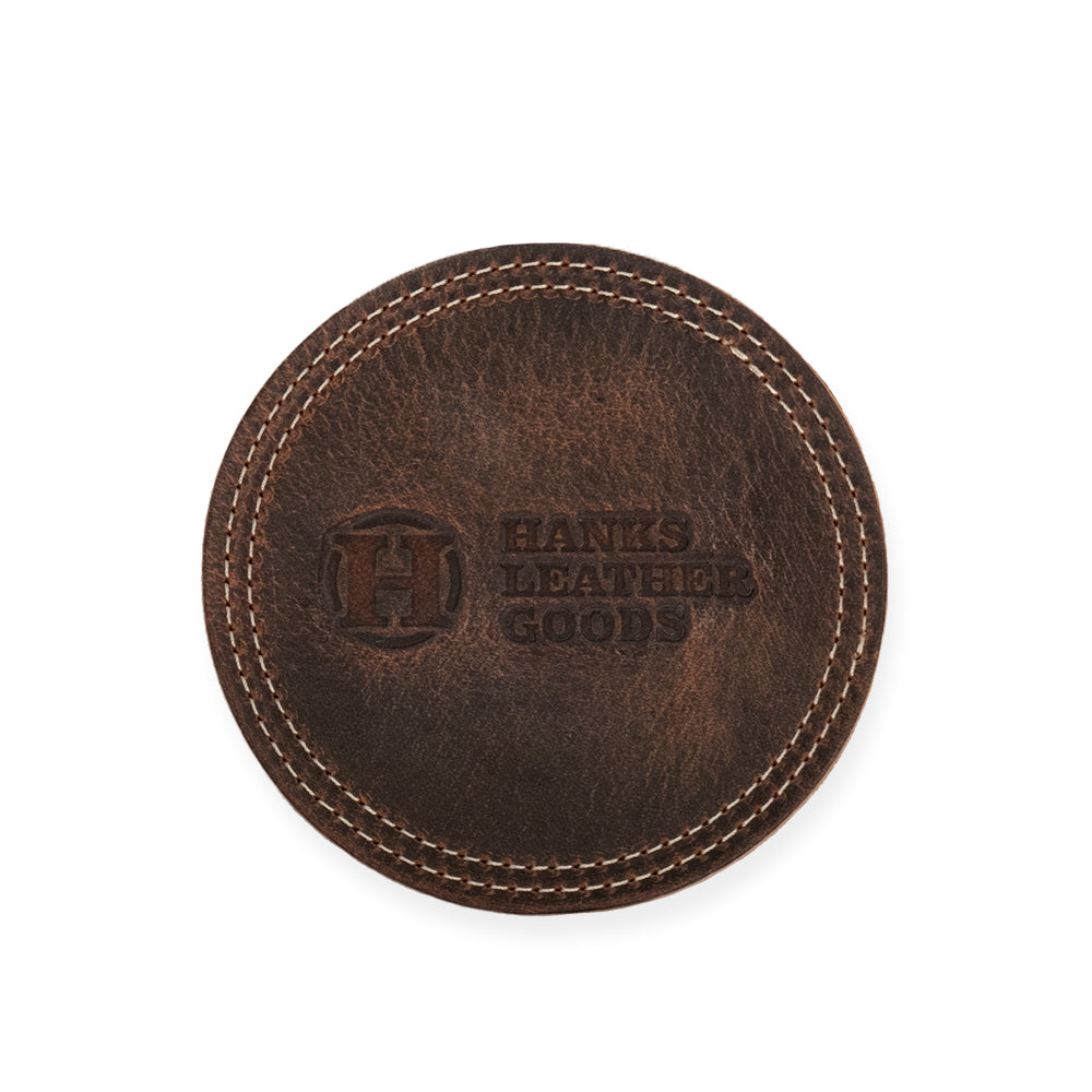 A round, brown leather coaster from Hanks Belts features distinct stitching along the edges and is embossed with the brand name in bold lettering.