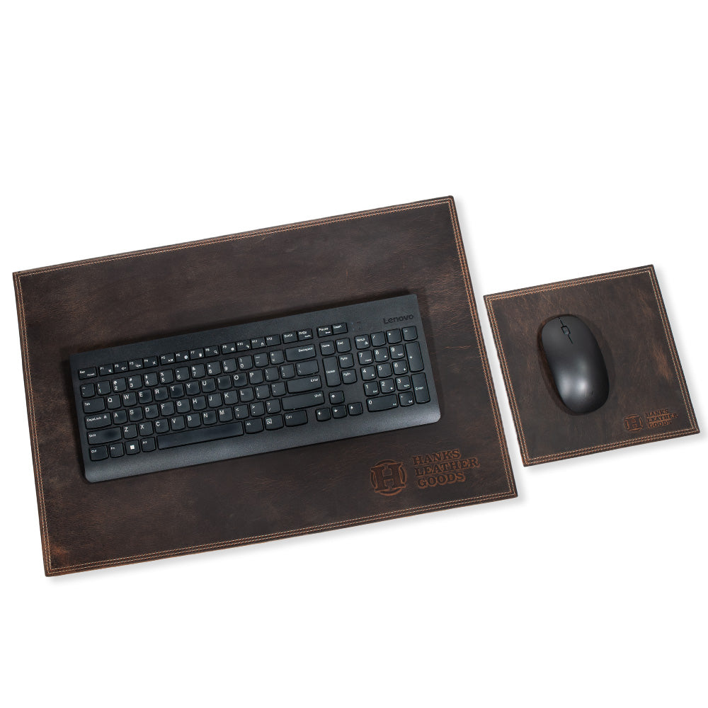 A black keyboard sits on a handmade, dark brown leather desk pad from Hanks Belts, proudly displaying the company logo. To its right is a matching North American Bison Leather mouse pad, also from Hanks Belts, with a black wireless mouse resting on top. The setup radiates a rustic and textured charm.