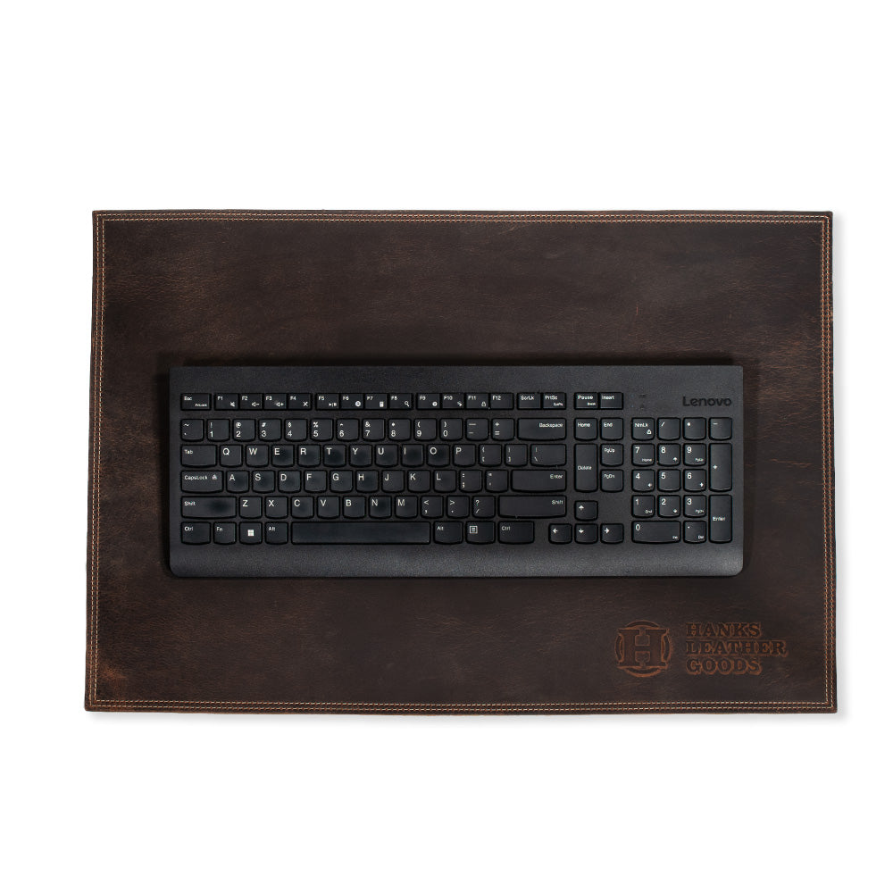 A black Lenovo keyboard sits atop a dark brown desk pad crafted from premium bison leather. This handmade piece, produced in the USA, showcases visible stitching and is embossed with &quot;Hanks Belts&quot; along with a decorative emblem in the bottom right corner.