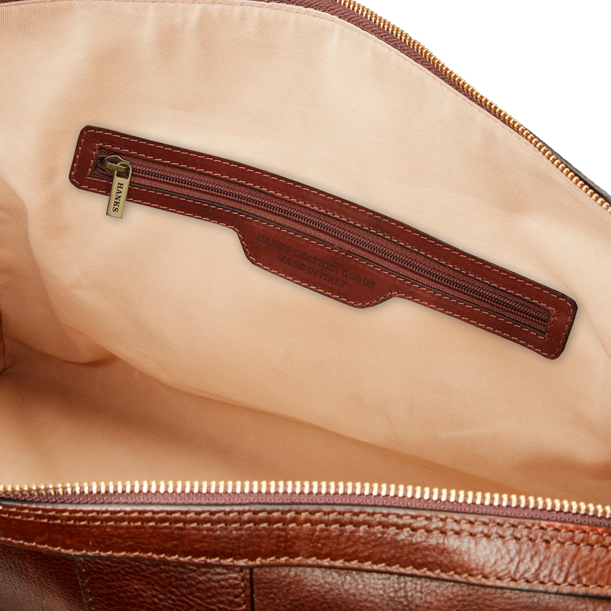 The Wanderer Duffel by Hanks is an open brown full-grain leather bag showcasing a beige interior lining with a zippered pocket, featuring a leather label embossed with text and a metallic zipper pull. Ideal for those who appreciate the exquisite details of an Italian-made luxury travel bag.