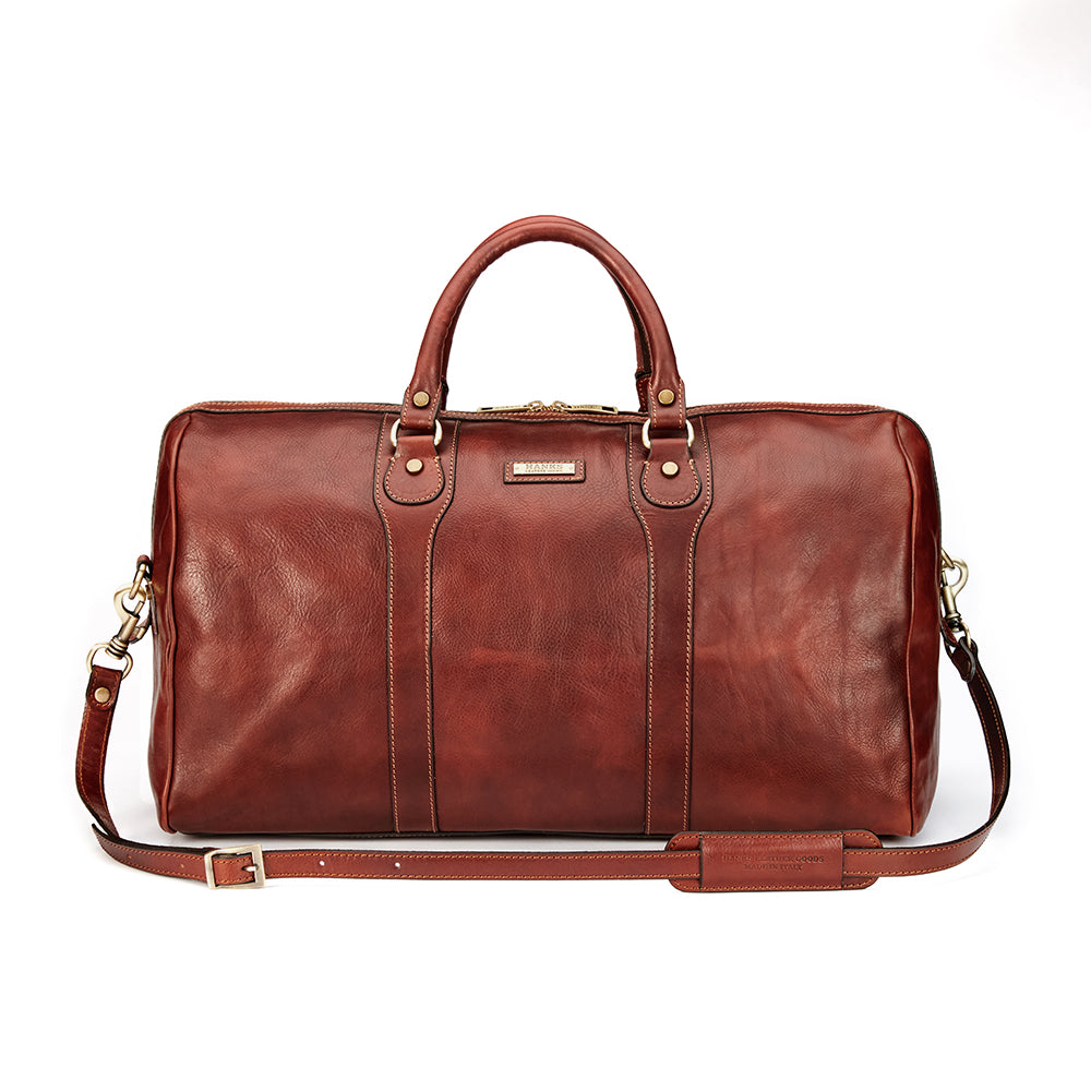 Introducing the Wanderer Duffel by Hanks, a luxurious travel companion crafted from full-grain leather in Italy. This exquisite brown duffel showcases dual handles and a detachable shoulder strap for versatile carry options. The front is elegantly adorned with a small metallic label, while two vertical seams add to its refined design.