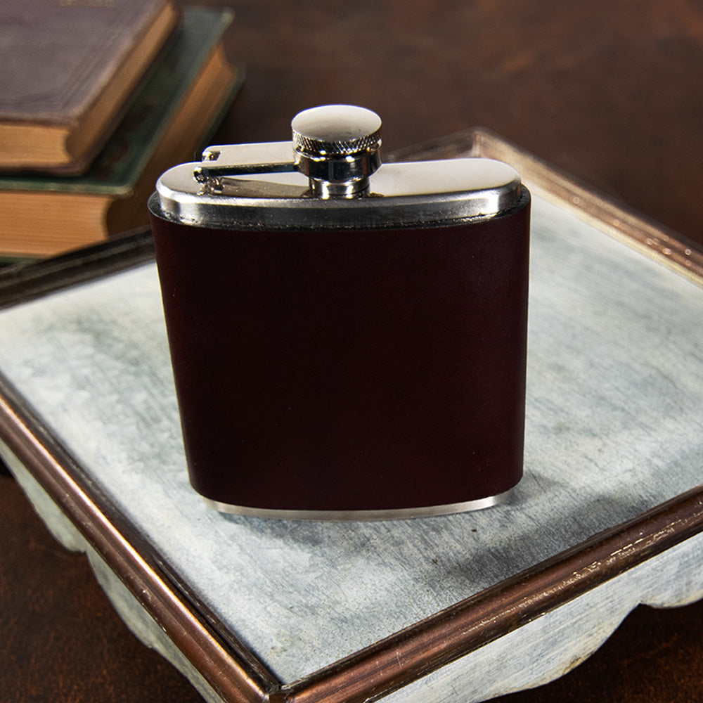 A compact Leather Covered Flask by Hanks Belts, featuring a dark finish and a shiny silver cap, rests on a white rectangular tray with raised edges. The tray is positioned on a brown surface. In the background, a couple of closed books are partially visible.