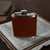 A Hanks Belts Leather Covered Flask, featuring USA-tanned brown leather and a metal cap, is placed on a white square tray. In the background, two stacked books with worn covers are partially visible.