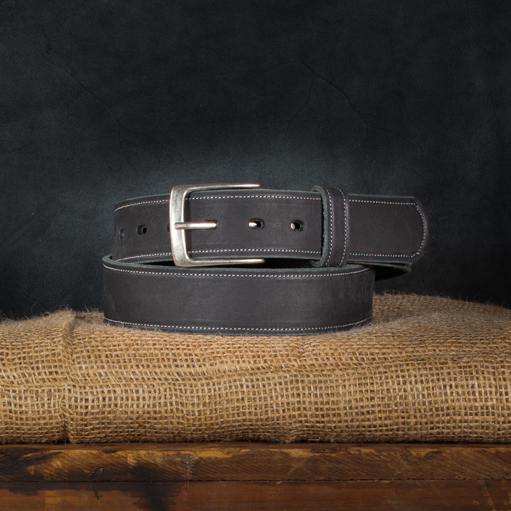 A neatly coiled Hanks Belts&#39; USA-made &quot;The Suede Belt - 1.5&quot; in dark leather with a silver buckle sits on burlap, set against a dark, textured background that highlights its smooth finish and white stitching.