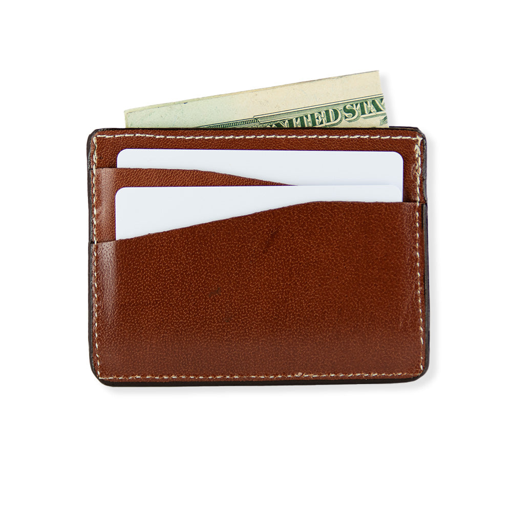 The Hanks Monroe Goatskin Card Holder, crafted from brown leather with white stitching and made in the USA, holds two white cards and a partially visible US dollar bill. The wallet is showcased on a white background.