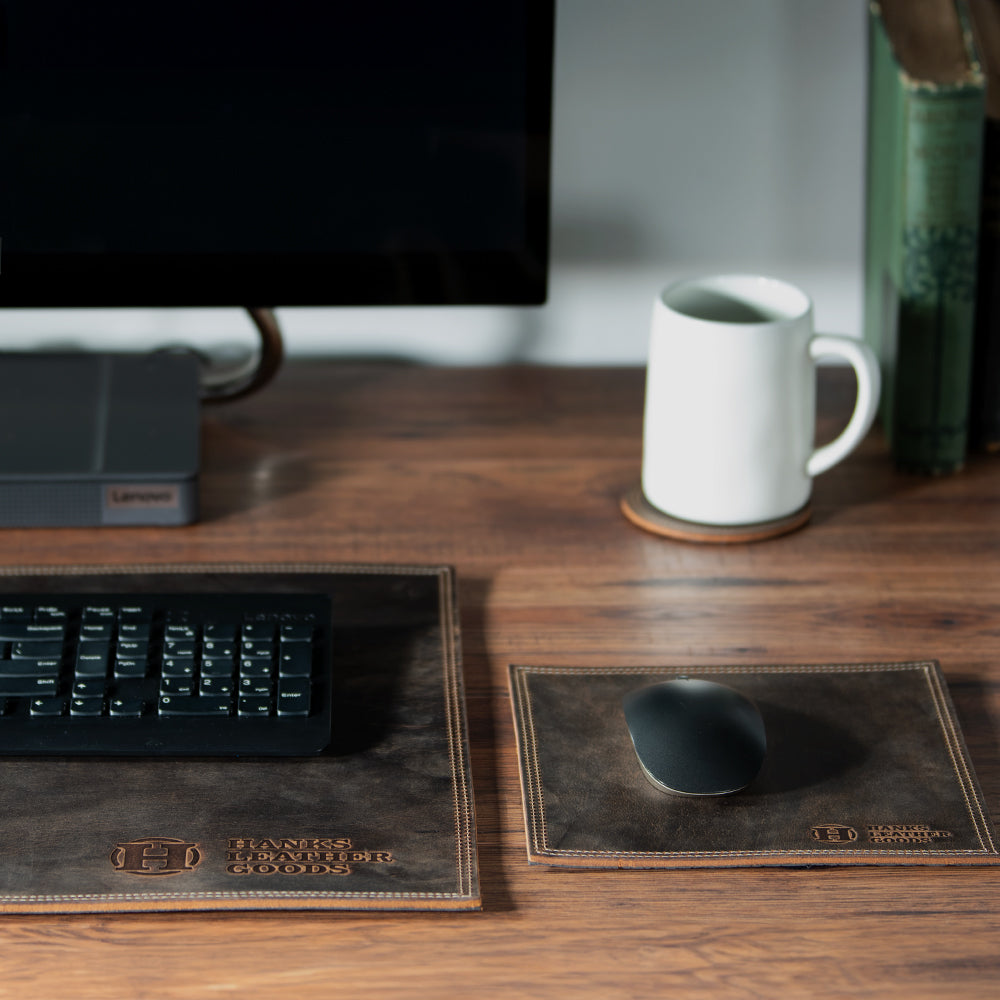 A Hanks Belts Desk &amp; Mouse Pad Set, featuring a computer keyboard and mouse on exquisite premium bison leather pads, is displayed on a wooden desk. A white mug enhances the aesthetic alongside a stack of books, with a computer monitor visible in the background. This arrangement radiates quality and craftsmanship, with each piece being meticulously handmade in the USA.