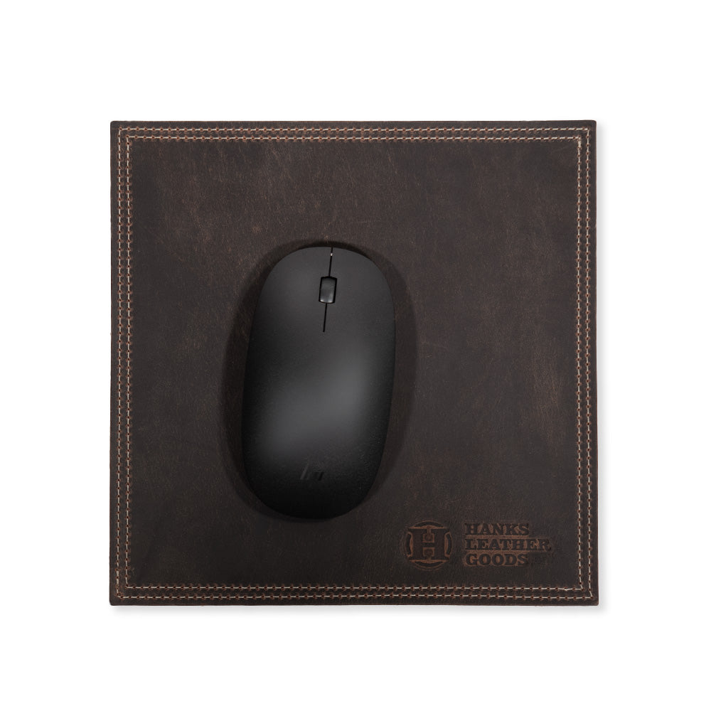 A black computer mouse rests on a dark brown bison leather mouse pad with decorative stitching, showcasing premium craftsmanship. This office accessory, branded by &quot;Hanks Belts,&quot; features their logo embossed in the bottom right corner.