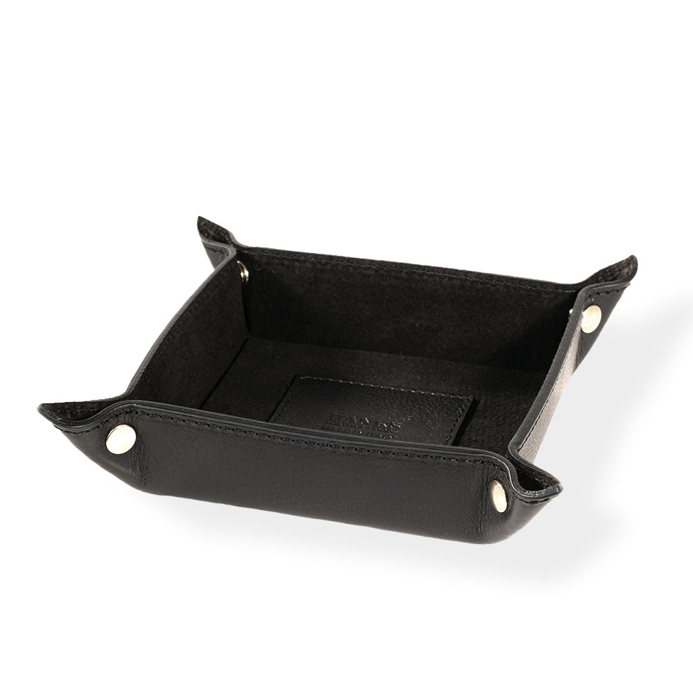 The Italian Compact Valet Tray by Hanks Belts is crafted from black Italian leather, featuring a compact rectangular shape with silver button snaps at each corner. The matching leather interior enhances its sleek look. Designed for travel and versatility, this tray is elegantly displayed on a plain white background.